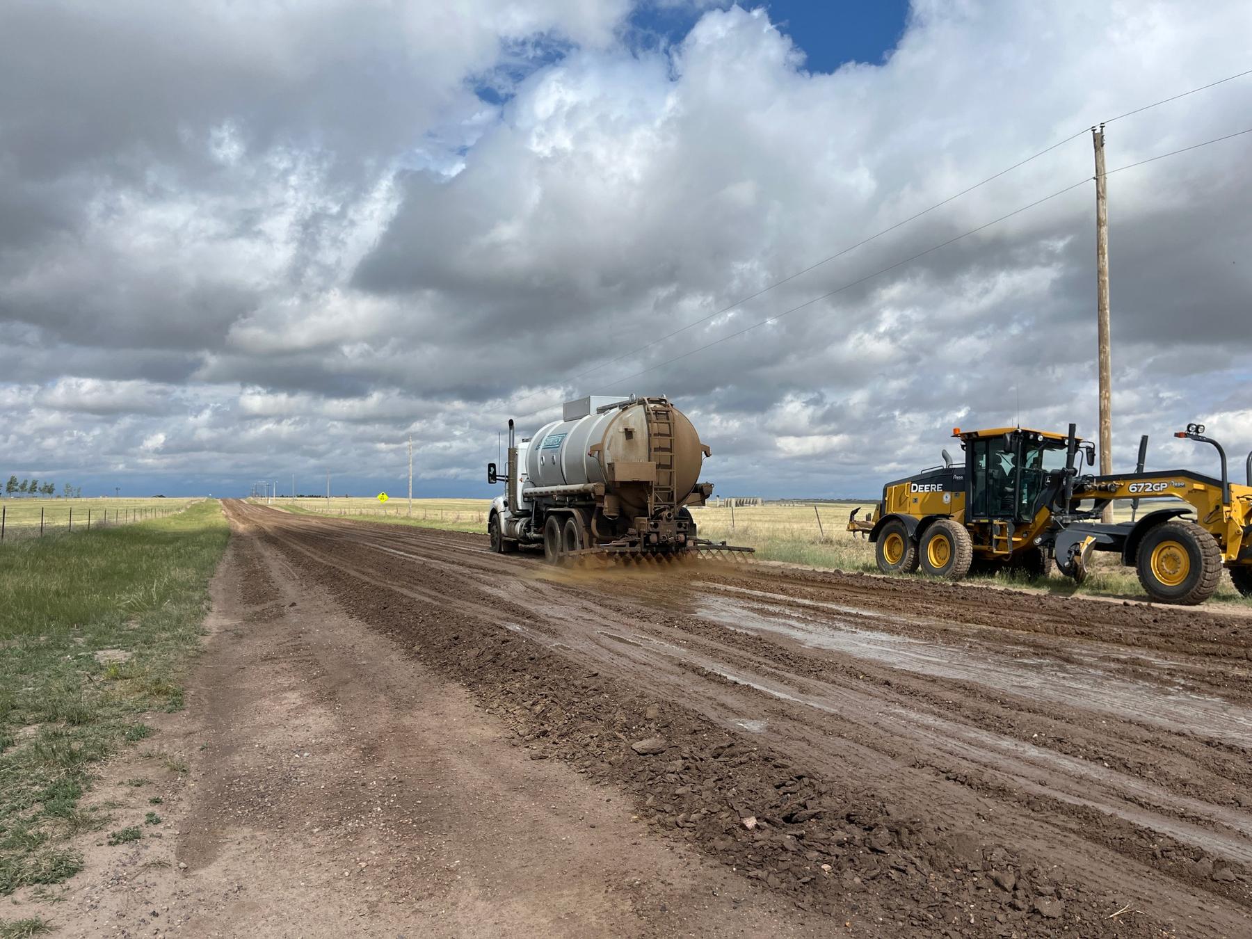 Tanker truck applying product behind a motor grader on a rural dirt road
